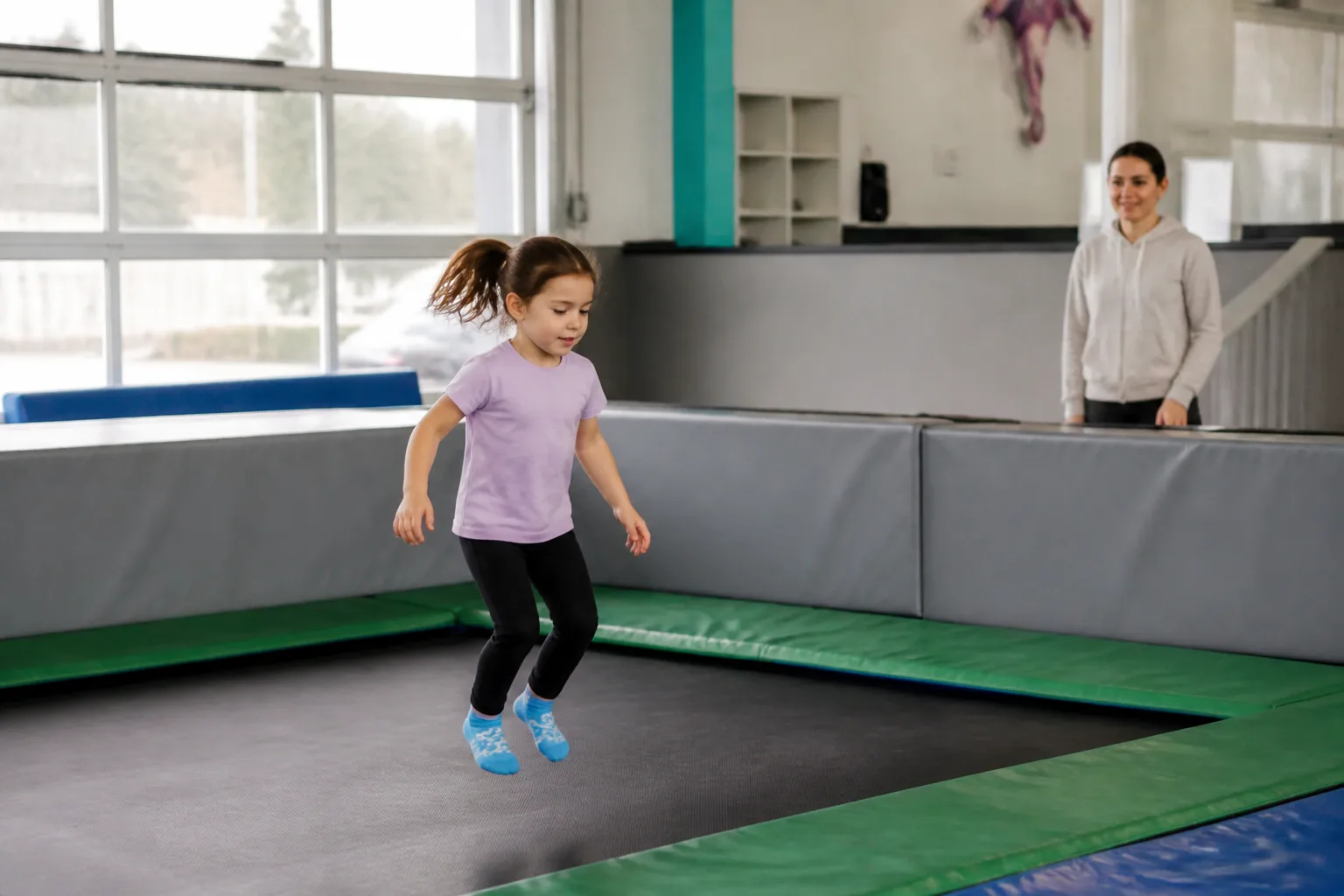 A child jumps on a trampoline with an adult supervising nearby.