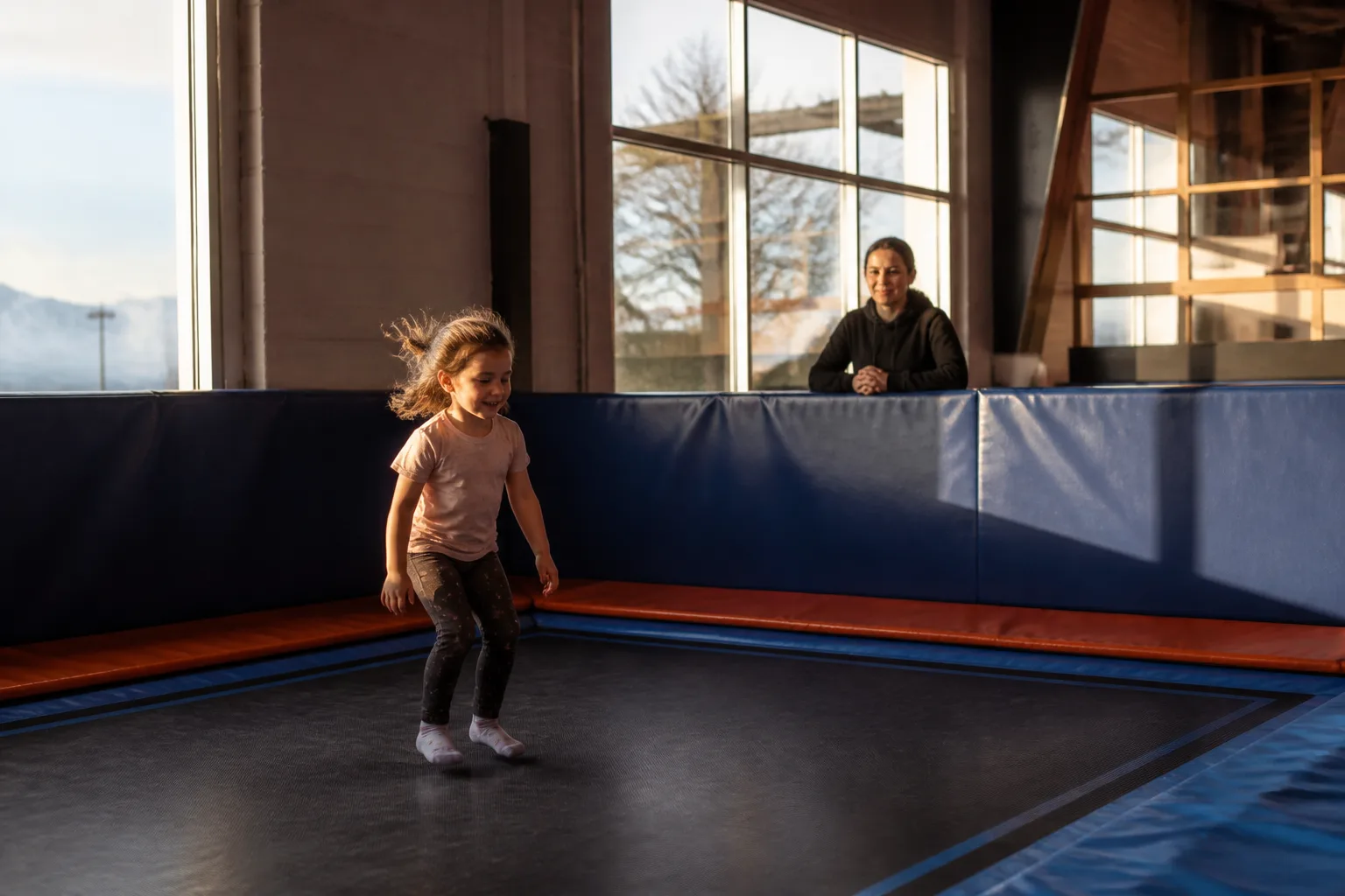A child bounces on a trampoline while an adult watches from behind padding.